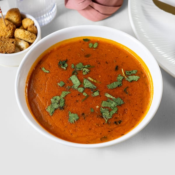 A bowl of vibrant orange Indian tomato soup, garnished with fresh green cilantro, next to croutons.