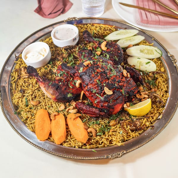 Pariwaar Delights' chicken biryani with roasted chicken, cashews, lemon, cucumber, and sauces on a silver platter.