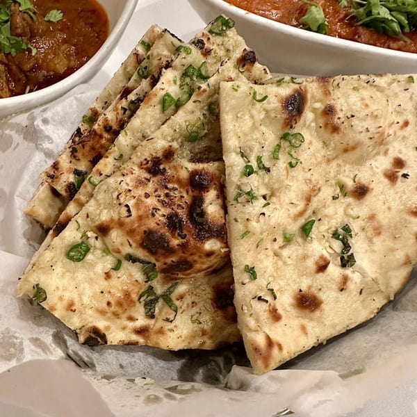 Close-up of garlic naan bread with green herbs, served with a bowl of curry in the background.