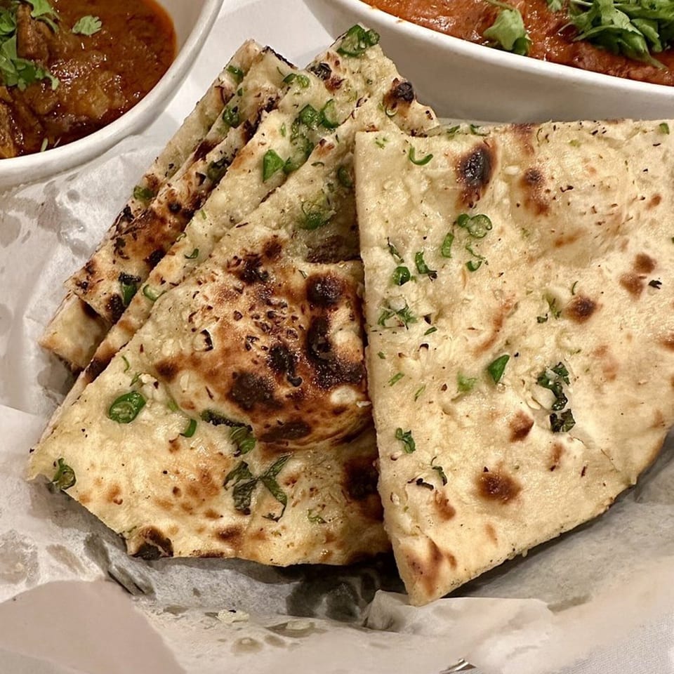 Close-up of garlic naan bread with green herbs, served with a bowl of curry in the background.