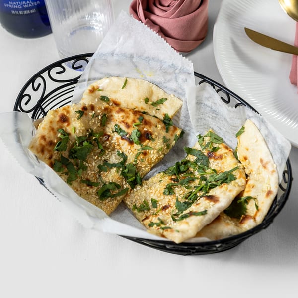 A black basket holds Indian naan bread, garnished with sesame seeds and fresh cilantro, on a white tablecloth.
