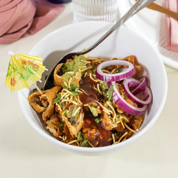 A bowl of Indian chaat with crispy fried pieces, green and brown sauces, red onions, and a tiny umbrella.