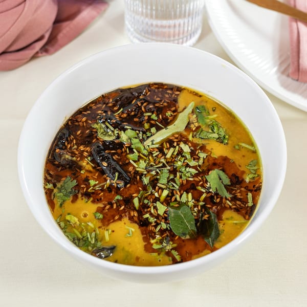 A white bowl of yellow Indian dal, topped with dark oil, cumin seeds, and fresh herbs.