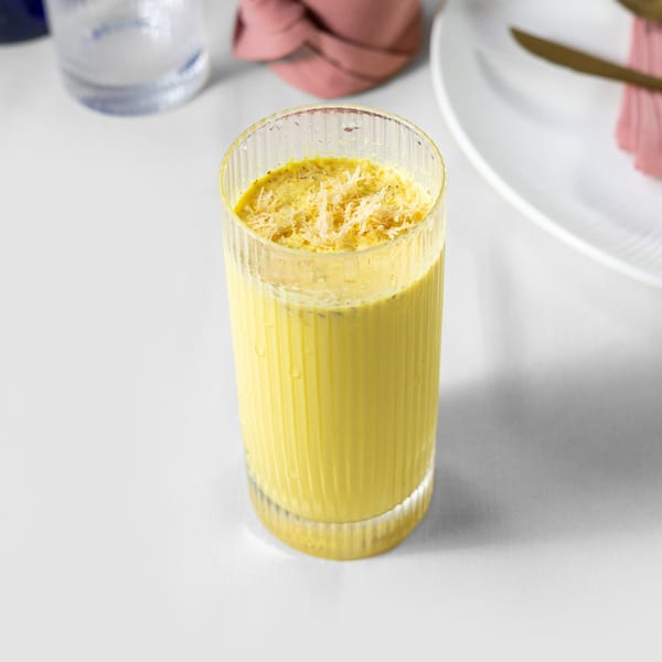 A tall glass of yellow Indian lassi, topped with shredded coconut, on a white table.